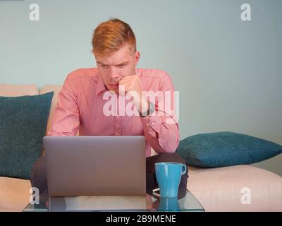 Un homme d'affaires blond caucasien travaille de la maison sur son ordinateur sur un canapé avec un fond bleu clair. Banque D'Images