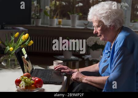 femme de 80 ans assise sur le canapé. Elle est en quarantaine. La femme senior écrit des messages avec sa petite-fille sur un ordinateur portable. Texte allemand. Banque D'Images