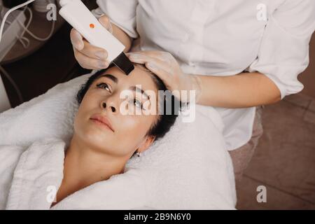 Soins de la peau du visage effectués au salon avec un appareil moderne par un jeune employé de spa Banque D'Images