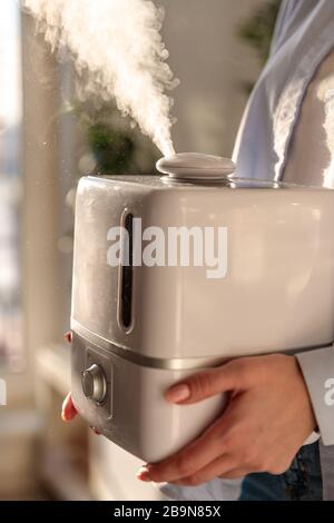 Femme tenant dans ses mains l'humidificateur d'huile d'arôme de travail dans la pièce ensoleillée. Augmentation de l'humidité de l'air à l'intérieur, conditions de vie confortables dans le chauffage Banque D'Images