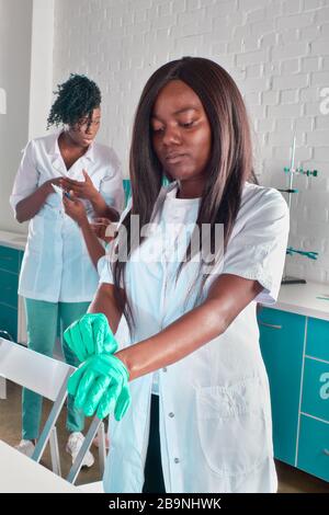 Le médecin, l'infirmière ou l'étudiant en médecine d'Afrique féminin en robe blanche protectrice met des gants médicaux. Jeune professionnel prêt à travailler à l'hôpital Banque D'Images