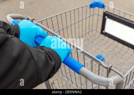 Une personne portant des gants en latex bleu pousse un panier vide dans un parking. Vue rapprochée centrée sur les mains. Banque D'Images