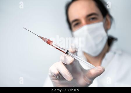 Tir sélectif de mise au point de la seringue avec le cadre central de l'aiguille rempli de liquide rouge, tenu par la main gantée de scientifique masqué hors de la mise au point sur un fond blanc Banque D'Images