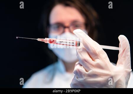 Dose sélective de mise au point de la seringue maintenue par le cadre central de la main avec gants remplie de liquide rouge. Derrière l'infirmière masquée hors de la mise au point sur fond noir Banque D'Images