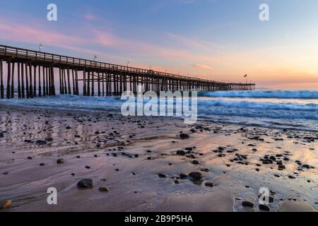 Jetée de Ventura au coucher du soleil, Ventura, Californie. Roches et sable en premier plan, l'eau s'est refluée dans la marée basse, avec des vagues entrantes. Banque D'Images