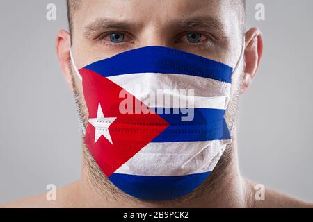 Jeune homme aux yeux douloureux dans un masque médical peint dans les couleurs du drapeau national de Cuba. Protection médicale contre les maladies aéroportées, coronavir Banque D'Images