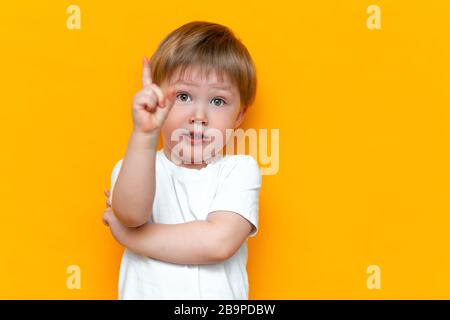 Portrait d'un joli petit garçon blond avec le doigt pointé vers le haut. Petit enfant en a l'idée. Enfant isolé sur fond noir jaune. Succès, idée brillante Banque D'Images