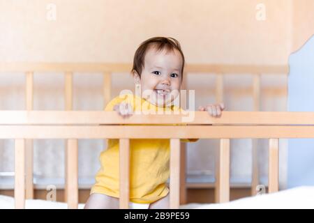 Bonne fille de bébé avec des cheveux sombres riant et souriant dans un t-shirt jaune mixte course kazakh et allemand debout dans lit bébé Banque D'Images