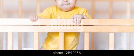 Jolie petite fille avec des cheveux foncés debout dans un lit de bébé à la maison dans un t-shirt jaune Banque D'Images