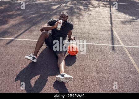 Activités en plein air. Homme africain assis sur le terrain de basket-ball avec de l'eau potable de balle souriant détendu Banque D'Images