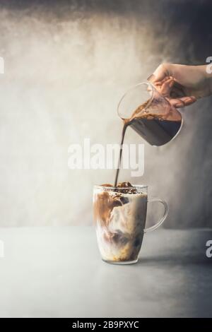 La main de barista qui verse de la crème à café dans une tasse en verre de Latte glacé sur une table rustique sur fond mural en béton. Cafetière glacée. Été rafraîchissant Banque D'Images