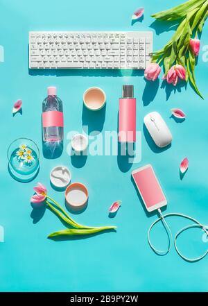 Produits cosmétiques avec maquette sur bureau turquoise. Clavier PC , smartphone avec écran vierge et fleurs de tulipes en fil et rose en lumière du soleil. Vue de dessus Banque D'Images