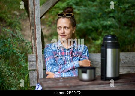 Jolie fille assise sur une table en bois avec thermos avec boisson chaude et chauffe les mains. La femme est froide et tente de se réchauffer. Voyage et a Banque D'Images