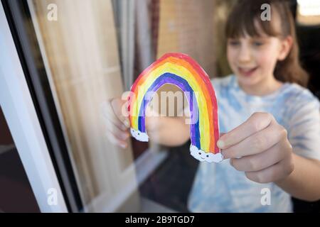 Fille mettant photo de Rainbow à la fenêtre à la maison pendant la pandémie de Coronavirus pour divertir les enfants Banque D'Images
