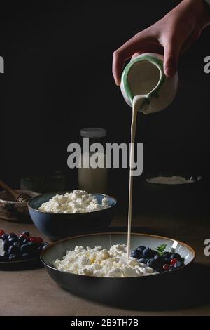 Produits laitiers pour le petit-déjeuner. Les mains du jeune homme versent de la crème de pichet au fromage cottage avec yaourt, lait, miel, bleuets dans un bol en céramique au-dessus de bro Banque D'Images