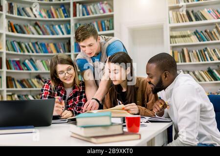 Quatre garçons et filles multiethniques souriants et positifs assis à la table dans une bibliothèque universitaire moderne et faisant conjointement des tâches à domicile ou des recherches d'informations Banque D'Images