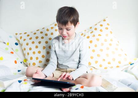 School Boy sur lit à la maison avec tablette numérique à la main, faire des devoirs. Formation à distance en ligne. Quarantaine Banque D'Images