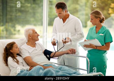 L'homme principal a sa tension artérielle prise alors que sa petite-fille se trouve à côté de lui dans un lit d'hôpital. Banque D'Images