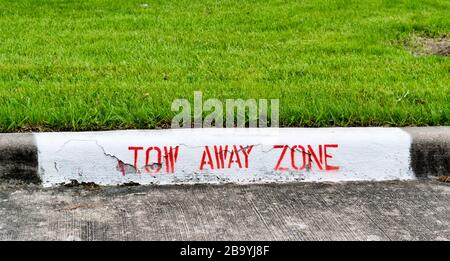 Panneau de zone de remorquage peint en lettres rouges sur un trottoir à travers avec une pelouse verte taillée derrière lui. Banque D'Images