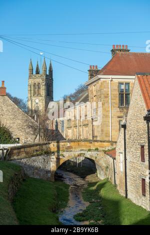 Belle vue sur Borough Beck, un ruisseau qui court à côté de Catlegate à Hemsley, avec un pont en pierre sur la beck et la tour de l'église de tous les Saints Banque D'Images