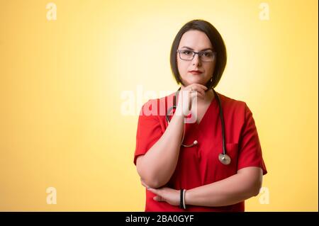 Portrait de belle femme médecin avec stéthoscope portant des gommages rouges, tenant la main sous son menton, ayant douté posant sur un dos isolé jaune Banque D'Images