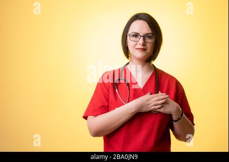 Portrait de belle femme médecin avec stéthoscope portant des gommages rouges, tient ses mains sur sa poitrine près de son coeur posant sur un backgr jaune isolé Banque D'Images