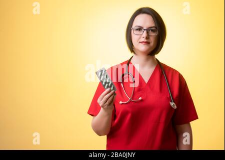 Portrait de belle femme médecin avec stéthoscope portant des gommages rouges, montrant des pilules vertes posant sur un dos isolé rose. Banque D'Images