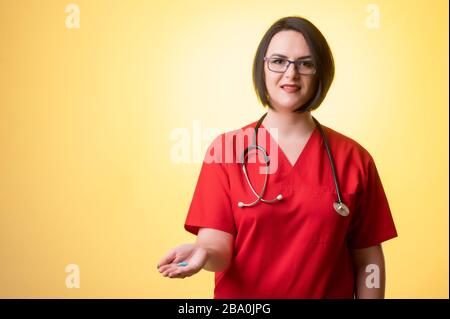 Portrait de belle femme médecin avec stéthoscope portant des gommages rouges, montrant des pilules bleues à la main posant sur un fond jaune isolé. Banque D'Images