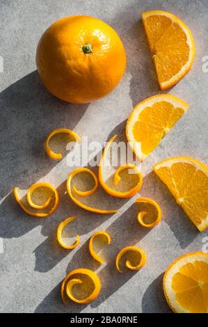 Fruits entiers orange et tranches de demi-cercle et boucles pelées de peau sur fond gris avec lumière du soleil Banque D'Images