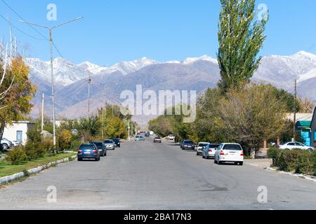 Rue asphaltée avec des voitures garées dans une petite ville près du lac Issyk Kul. La chaîne des montagnes enneigées et les arbres visibles à Balykchy, au Kirghizstan. Banque D'Images