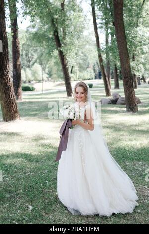 Magnifique jeune mariée en robe blanche tenant bouquet dans le parc Banque D'Images