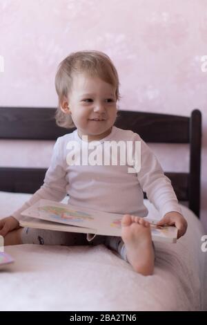 Un petit garçon aux cheveux blonds est assis sur le lit et souriant, tenant un livre dans ses mains. Un petit garçon est assis sur un lit avec un livre dans ses mains pendant Banque D'Images