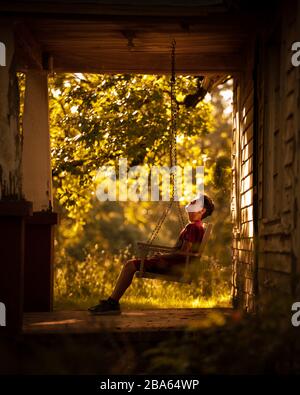 Un jeune garçon est assis sur un porche de campagne balancent à explorer une ferme abandonnée dans les montagnes Ozark. Banque D'Images