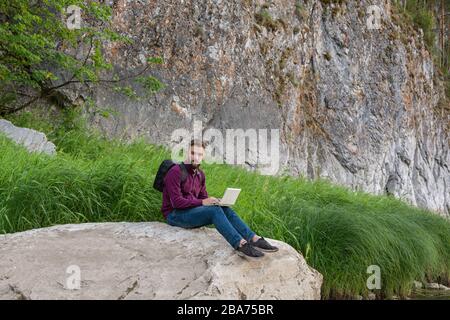 Jeune homme voyageur avec sac à dos se trouve sur un gros rocher plat dans un endroit pittoresque près du mur de montagne travaillant sur ordinateur portable en beau temps ensoleillé. Vue latérale. Travail Banque D'Images