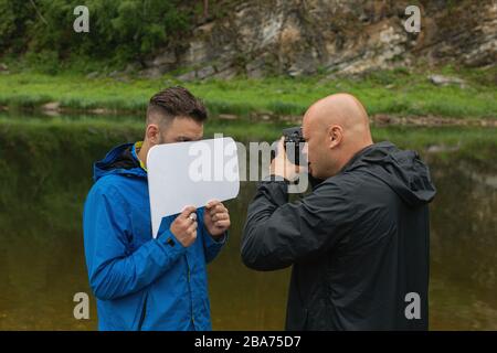 Travail de photographe avec modèle en extérieur. Photographe prenant gros plan de jeune homme beau debout sur la rive de la rivière cachant une partie de son visage beh Banque D'Images