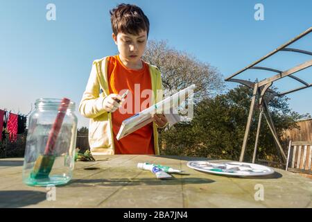 Un jeune garçon qui fait une peinture sur une table dans le jardin à l'arrière une journée ensoleillée. Banque D'Images