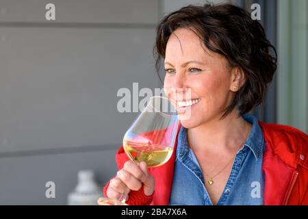 Femme brunette avec des cheveux courts, portant une chemise denim bleue et une veste rouge, tenant un verre de vin blanc, souriant et regardant de côté. Portrait buste avec Banque D'Images