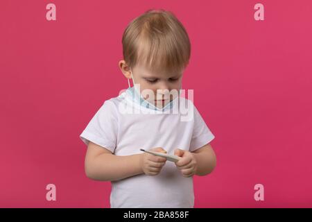 Un jeune médecin enfant avec un masque médical tient un thermomètre dans ses mains et regarde la température. Gros plan studio tourné sur un dos rose brillant Banque D'Images