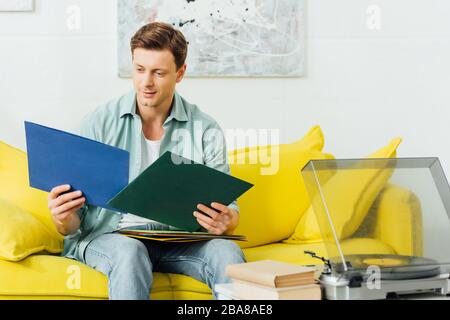 Beau homme tenant des disques vinyles près du lecteur de disques et des livres sur la table de café Banque D'Images