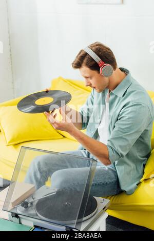 Vue latérale du beau homme dans un casque qui tient un disque vinyle près des livres et un lecteur de disques sur la table basse Banque D'Images