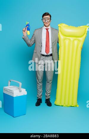 jeune homme d'affaires dans un masque de plongée avec matelas gonflable et pistolet à eau tout en se tenant près d'un réfrigérateur portable sur fond bleu Banque D'Images