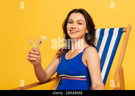 une femme gaie en maillot de bain bleu assis dans une chaise longue avec un verre de cocktail et regardant un appareil photo isolé sur le jaune Banque D'Images