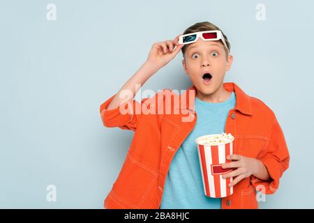 enfant choqué avec des lunettes en relief tenant le seau avec le pop-corn sur fond bleu Banque D'Images