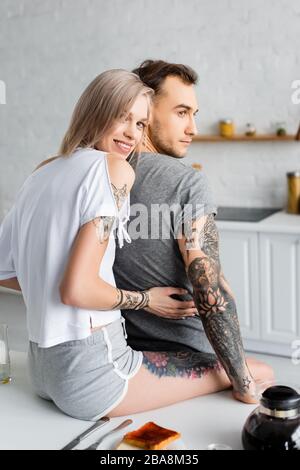 Belle fille souriante à l'appareil photo tout en embrassant tatoué petit-déjeuner dans la cuisine Banque D'Images