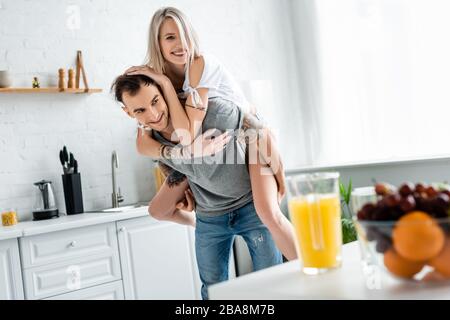 Foyer sélectif de la fille souriante piggybacking sur tatoued petit ami près des fruits et jus d'orange sur la table de cuisine Banque D'Images