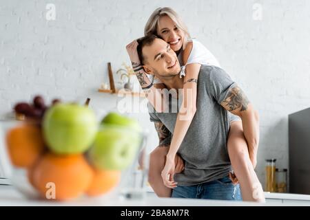 Foyer sélectif de la fille souriante piggybacking sur tatoued petit ami près de bol de fruits frais sur table de cuisine Banque D'Images