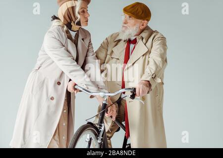 Couple senior élégant regardant l'un l'autre près du vélo isolé sur gris Banque D'Images