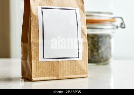 Sac en papier kraft marron avec étiquette blanche vierge devant un bocal en verre à balles et joint en caoutchouc orange avec herbes séchées ou thé sur fond blanc. Banque D'Images