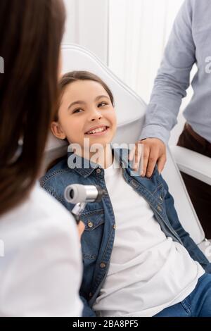 vue courte du père touchant l'épaule de la fille assise dans la chaise médicale près de l'otoscope du médecin Banque D'Images