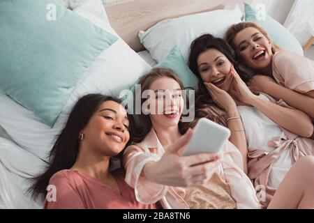 Vue panoramique sur les femmes multiculturelles excitées prenant le selfie tout en se reposant sur un lit à la fête de Bachelorette Banque D'Images